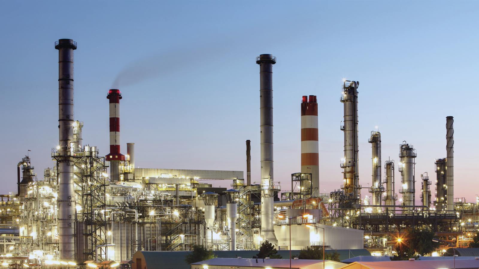Refinaria iluminada ao entardecer, com chaminés e torres de processo